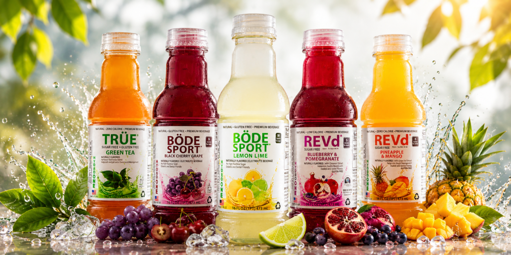 Six colorful beverage bottles displayed in a row with fruit and ice around, on a bright outdoor setup.